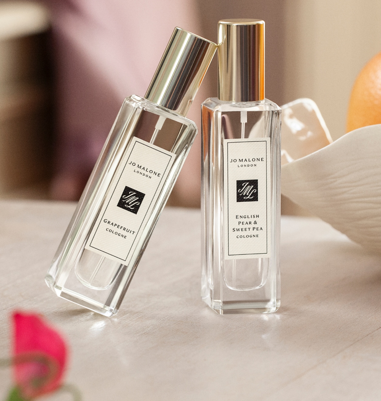 Two Jo Malone London perfume bottles with gold caps on a table, set beside a bowl of orangesw with small pink flowers in front.