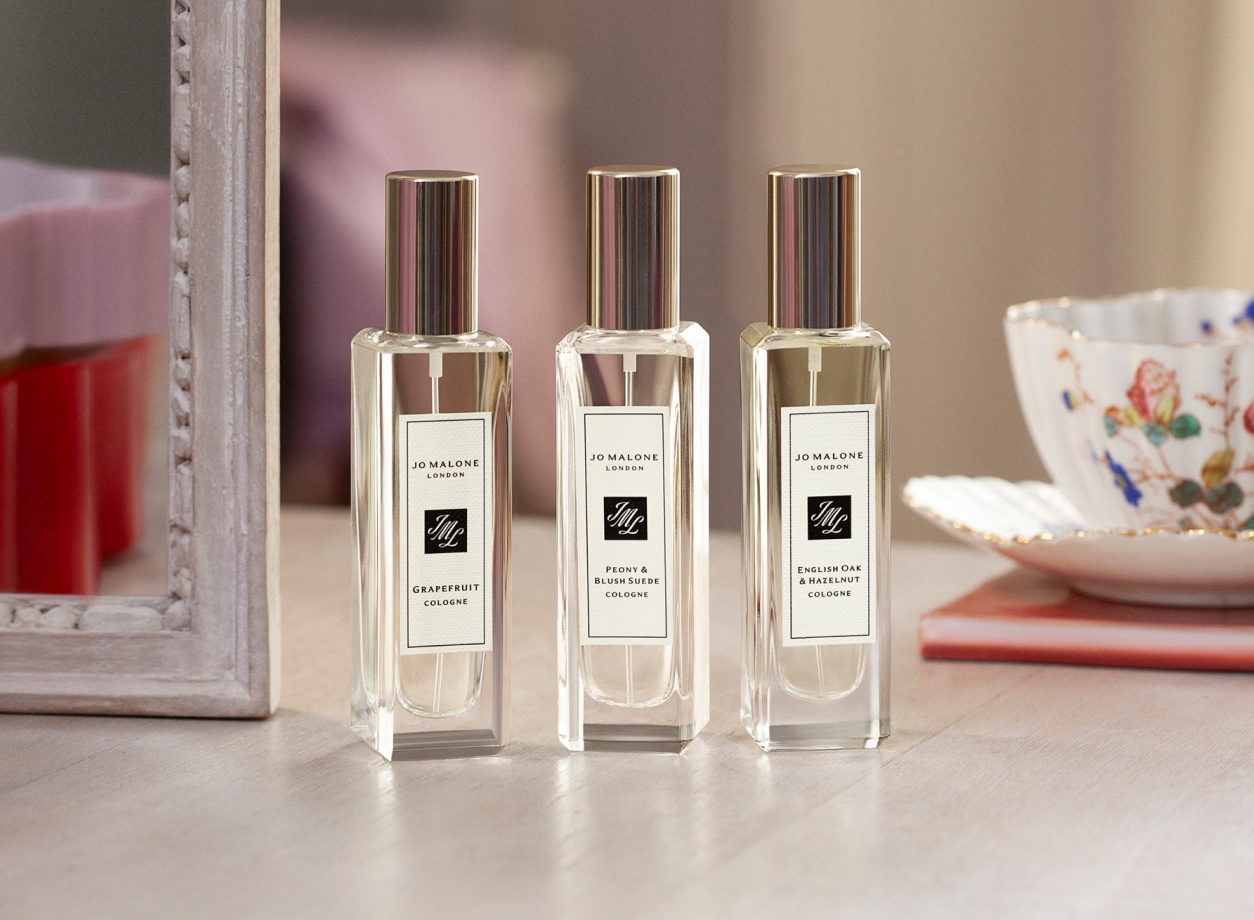 Three Jo Malone London perfume bottles with silver caps on a wooden table, set beside a decorative teacup and a framed mirror